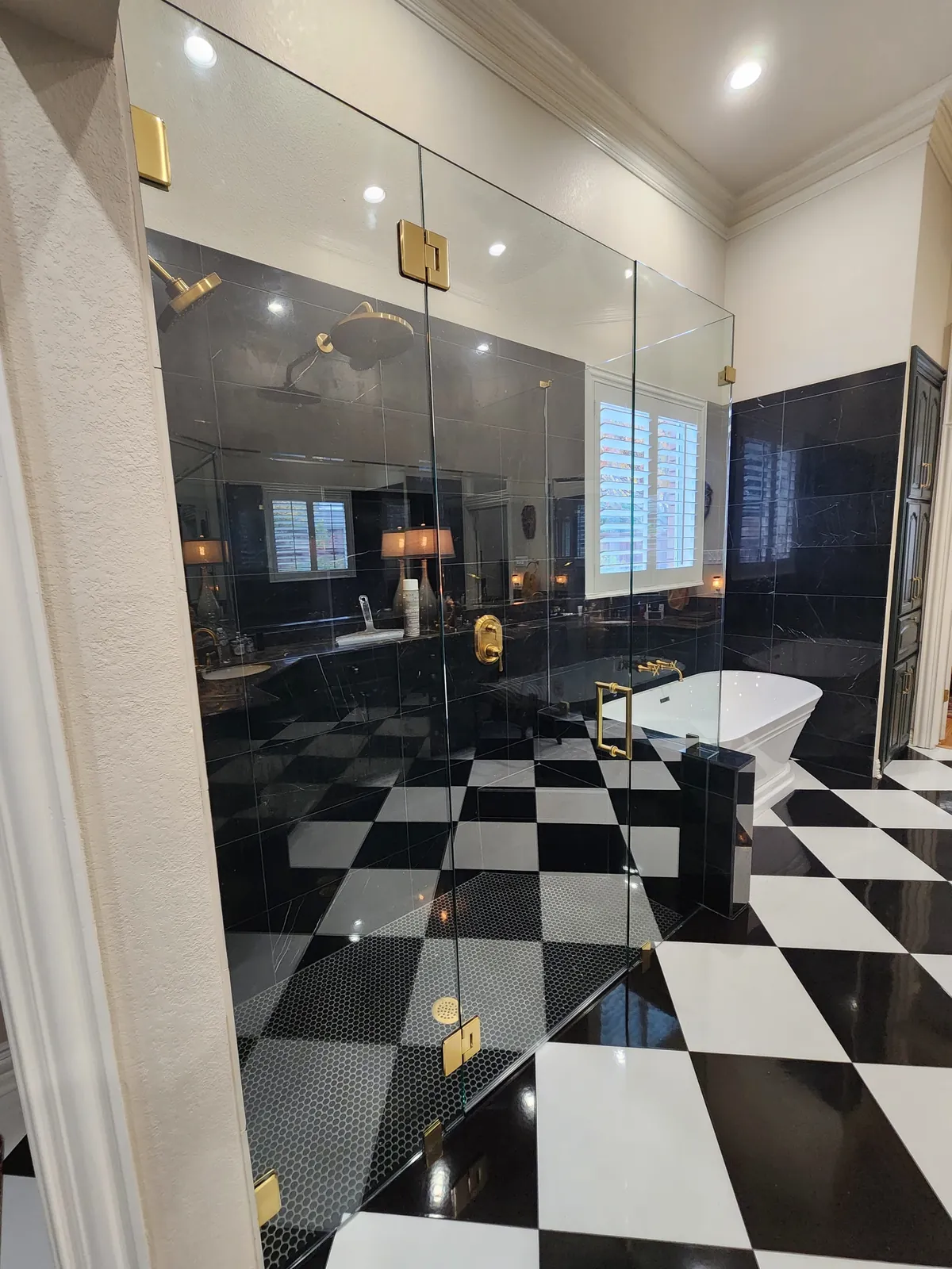 Full-height frameless glass enclosure with brass hinges and handle beside a black and white checkerboard floor