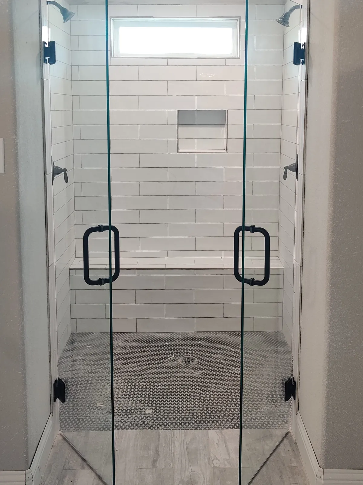 Pair of frameless glass shower doors with matte black pulls opening into a white subway-tile shower