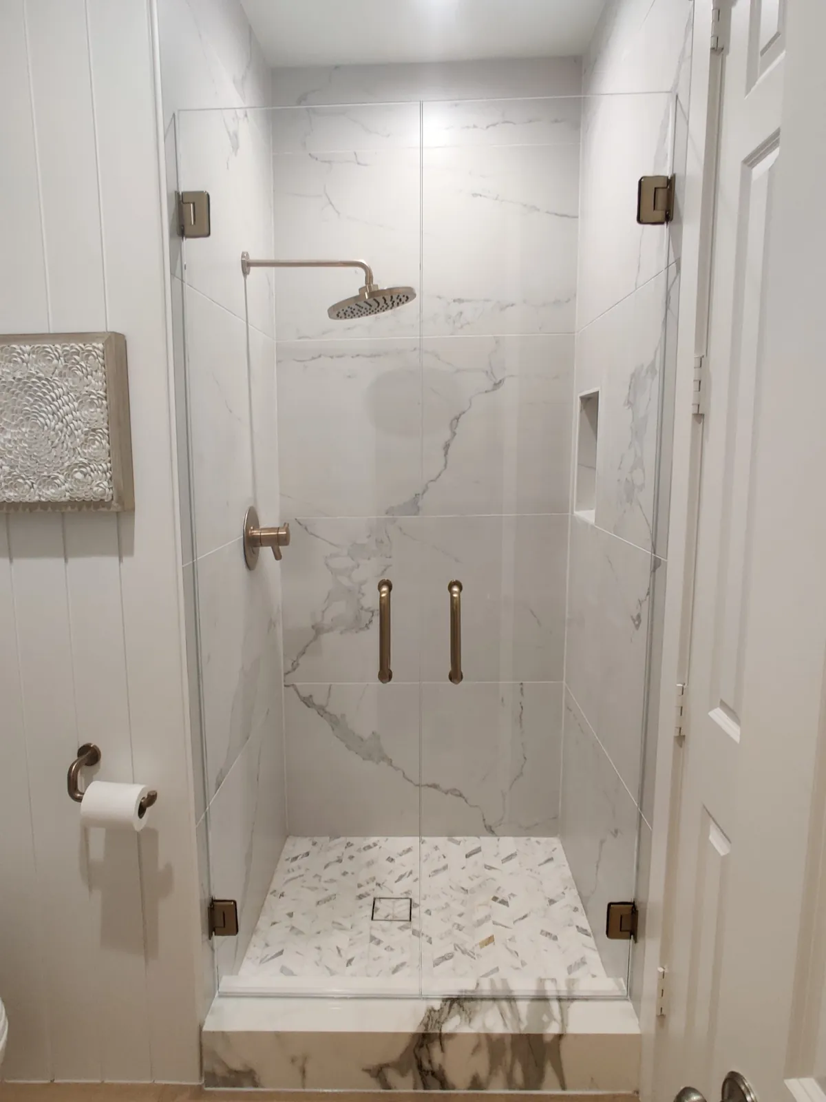 Pair of frameless glass shower doors with brushed bronze hinges and pulls in a white marble-look shower