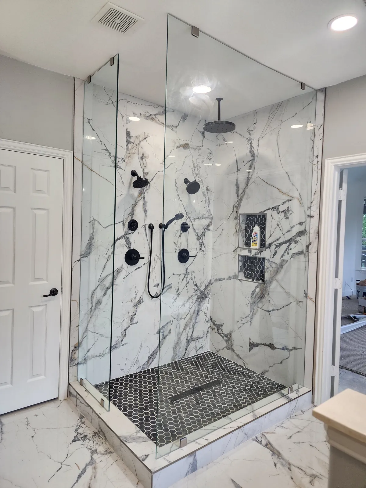 Frameless glass shower enclosure with matte black fixtures, white marble-look walls and a black hex tile floor