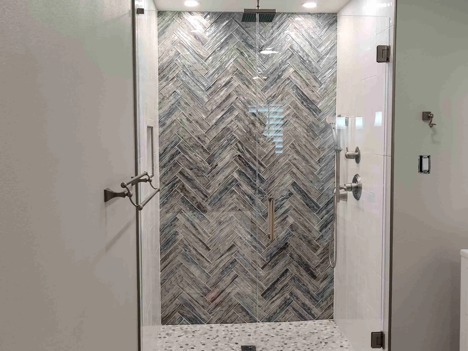 Frameless glass shower door and fixed panel in front of a blue-gray herringbone accent wall and pebble floor