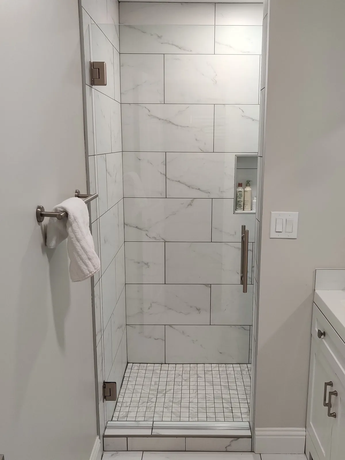 Frameless glass shower door with brushed nickel hinges and a towel bar, white marble-look tile inside