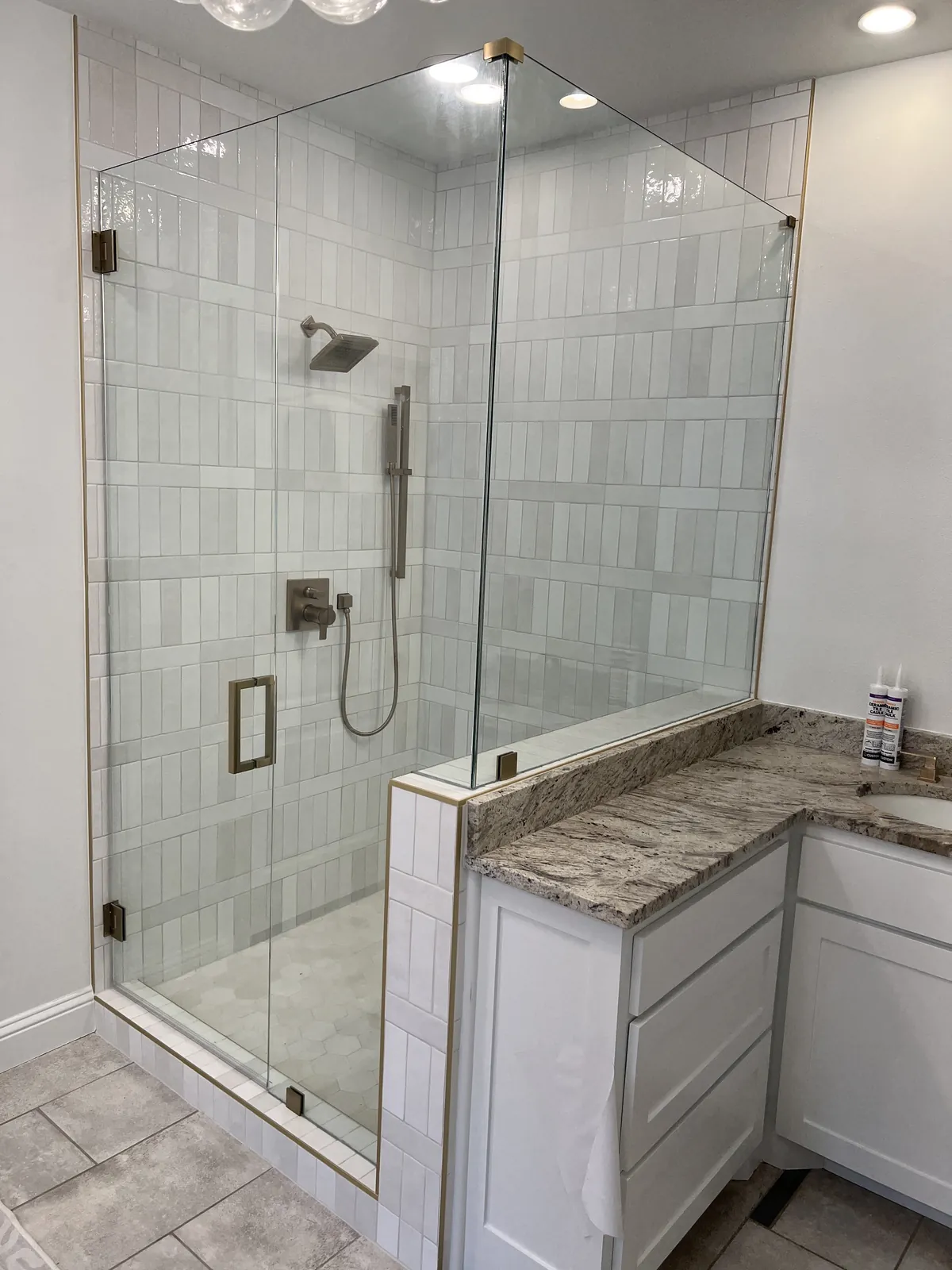 Frameless glass door and panels on a tiled knee wall with brass hinges, beside a granite-topped vanity