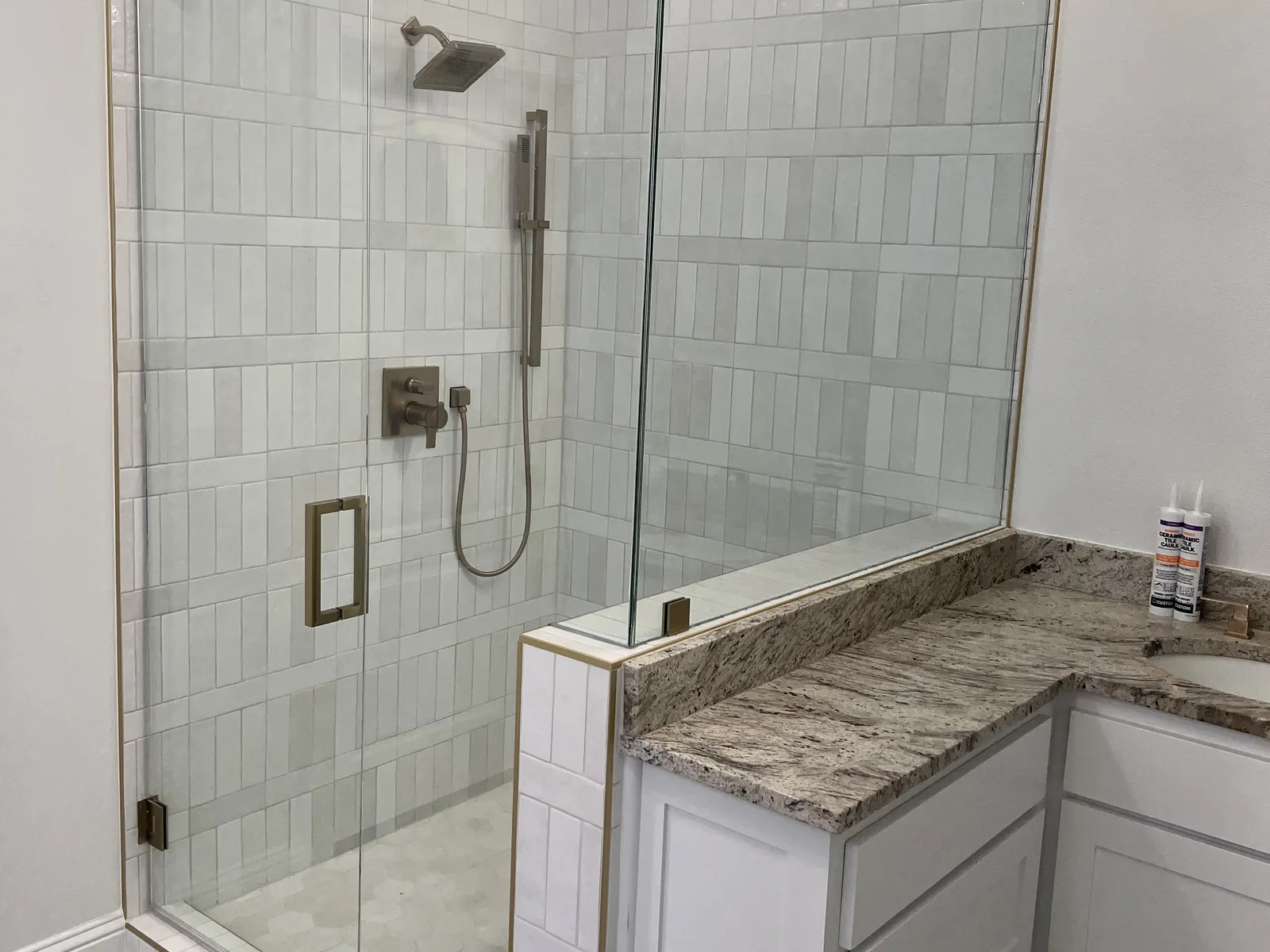 Frameless glass door and panels on a tiled knee wall with brass hinges, beside a granite-topped vanity