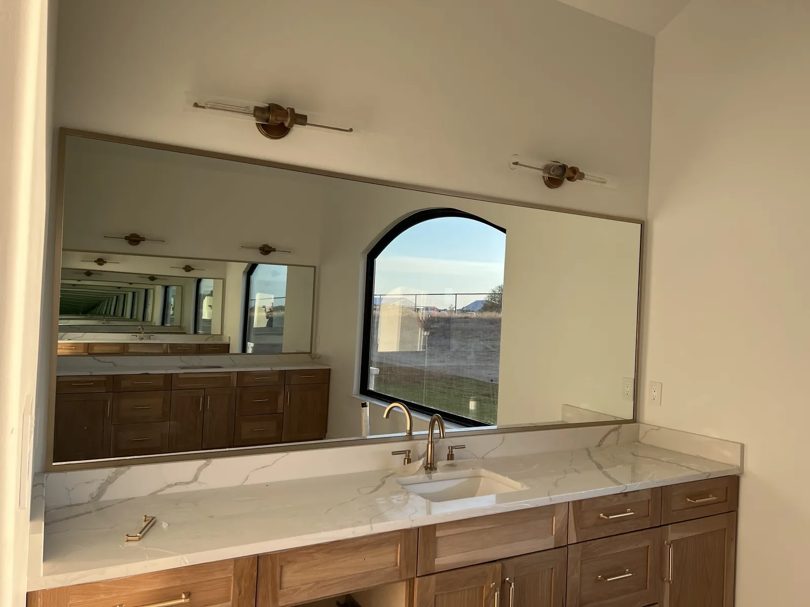 Long thin-framed mirror above a wood vanity, reflecting an arched window