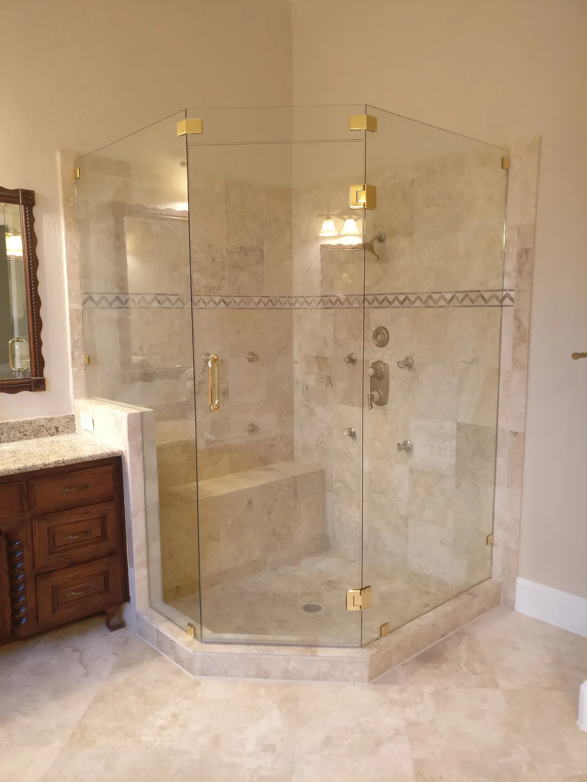 Neo-angle frameless glass shower with brass hardware, travertine-look tile and a built-in bench