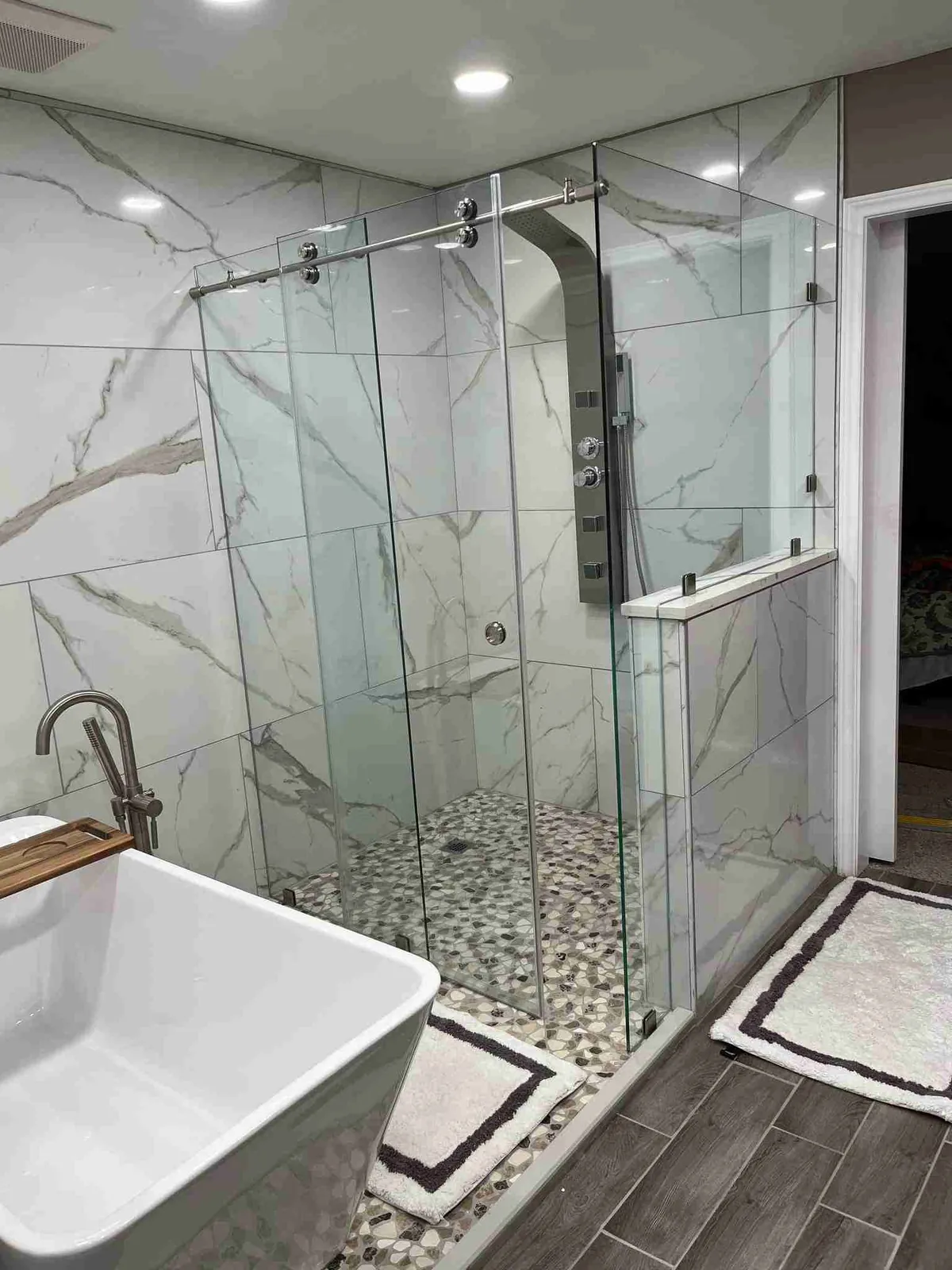 Frameless glass shower with a sliding door on a rail and a fixed panel on a knee wall, beside a freestanding tub